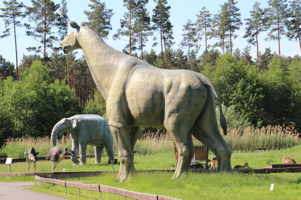 Large dinosaur statues in a lush park with a sunny backdrop. A tall sauropod stands prominently, surrounded by trees and grass, evoking prehistoric times.