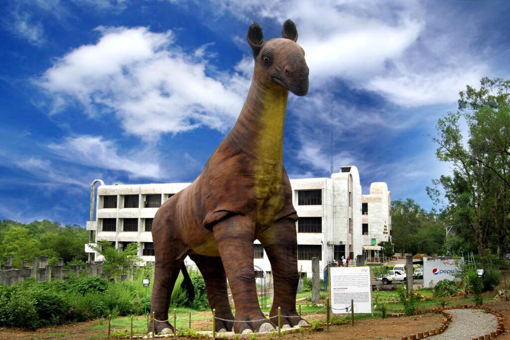 A large dinosaur statue stands prominently in front of a white building, surrounded by greenery under a bright, cloudy blue sky, conveying a sense of awe and history.
