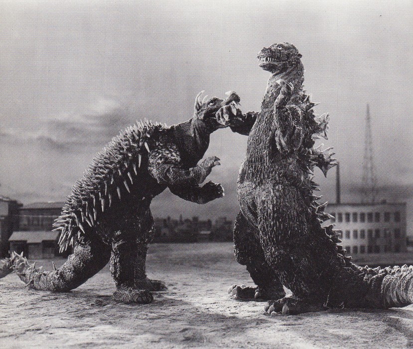 Two towering, spiky monsters face off dramatically against a city backdrop under a cloudy sky, evoking tension and impending conflict.