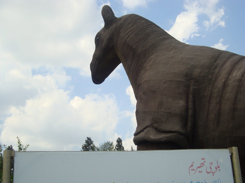 Large camel statue viewed from the side against a cloudy sky. A sign with text in a local script is partially visible at the bottom. Calm, majestic setting.