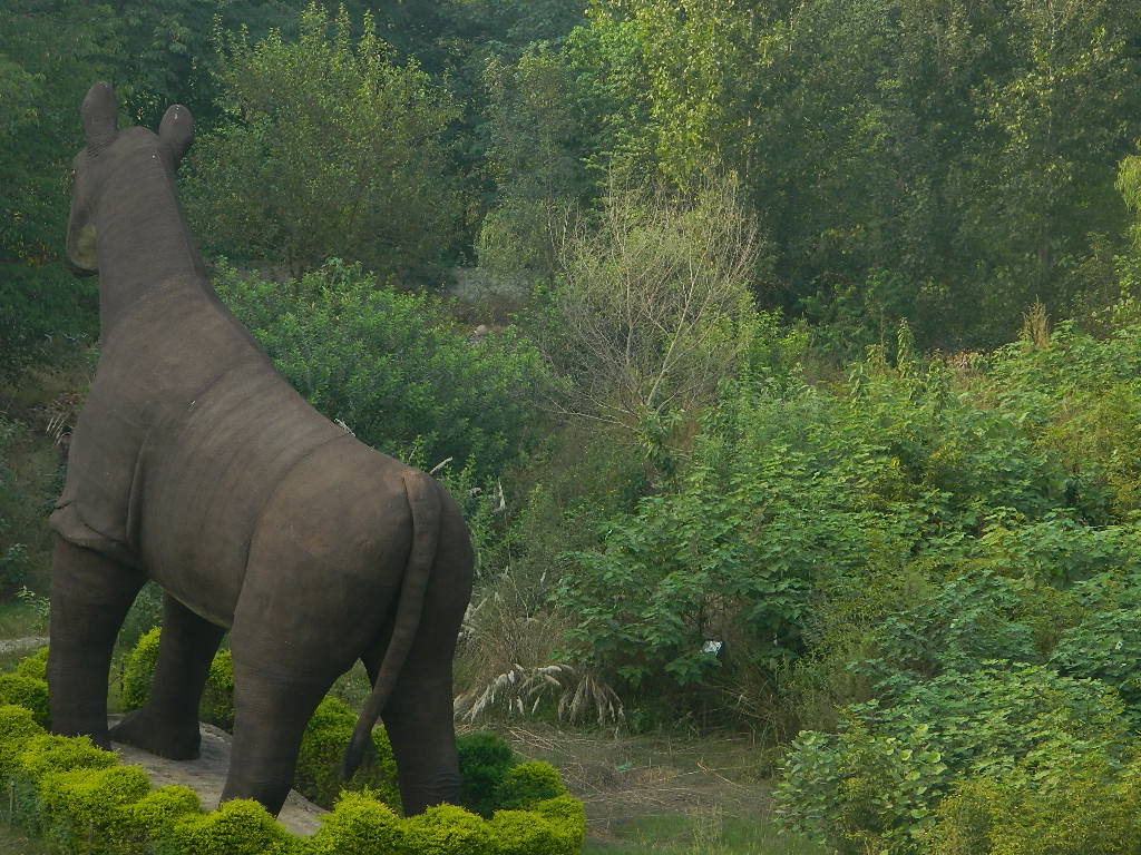 A large brown statue of an animal resembling a horse stands among lush green foliage. The scene is peaceful, surrounded by dense, vibrant trees.