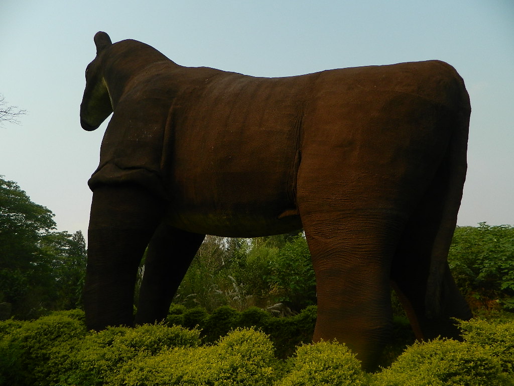 A large, brown elephant sculpture made of straw and grass stands on lush, green bushes. The sky is clear and pale, creating a serene atmosphere.