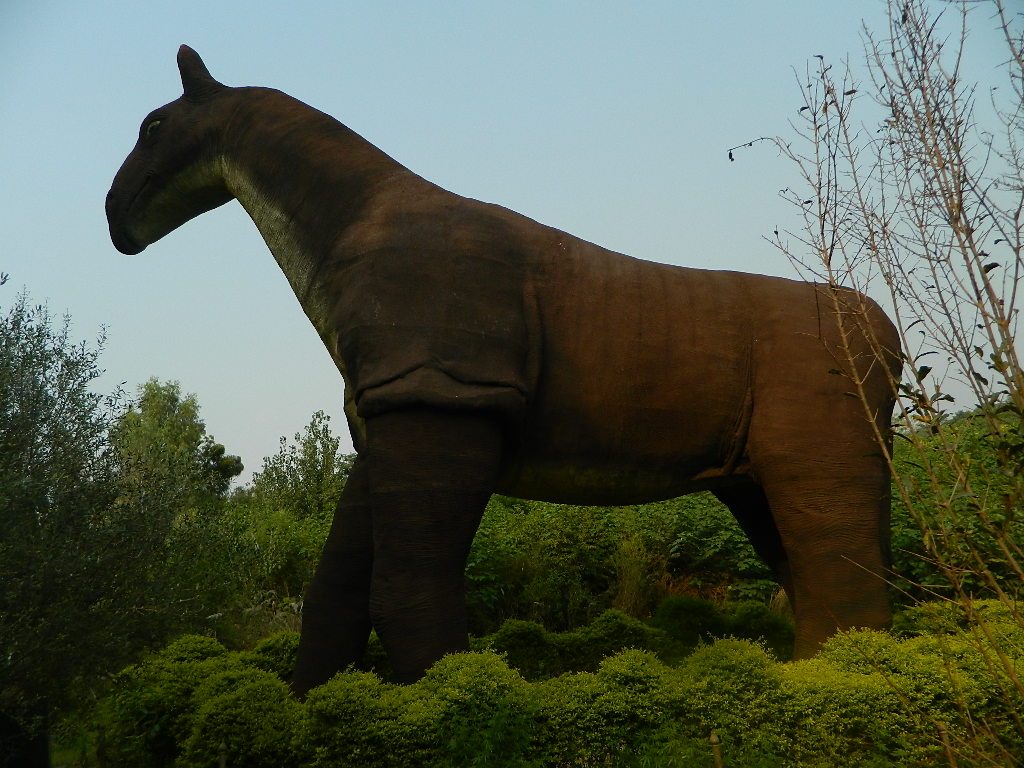 A large equine sculpture stands surrounded by lush greenery against a clear blue sky. Its dark brown color contrasts with the vibrant foliage, conveying grandeur.