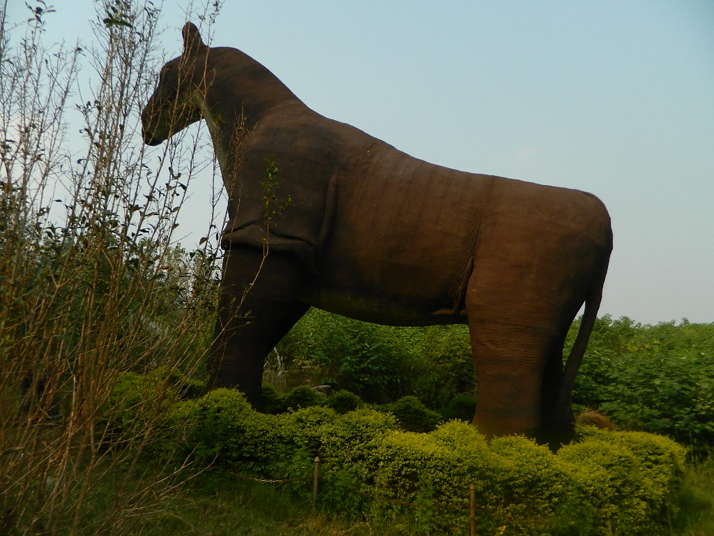 A large, brown T-Rex sculpture stands among green bushes and tall plants, blending into a natural setting under an overcast sky. The tone is tranquil and rustic.