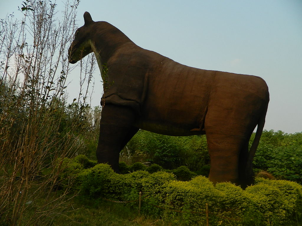 Large, brown, elephant-like statue stands among lush greenery with a clear sky. The scene conveys a tranquil and natural atmosphere.