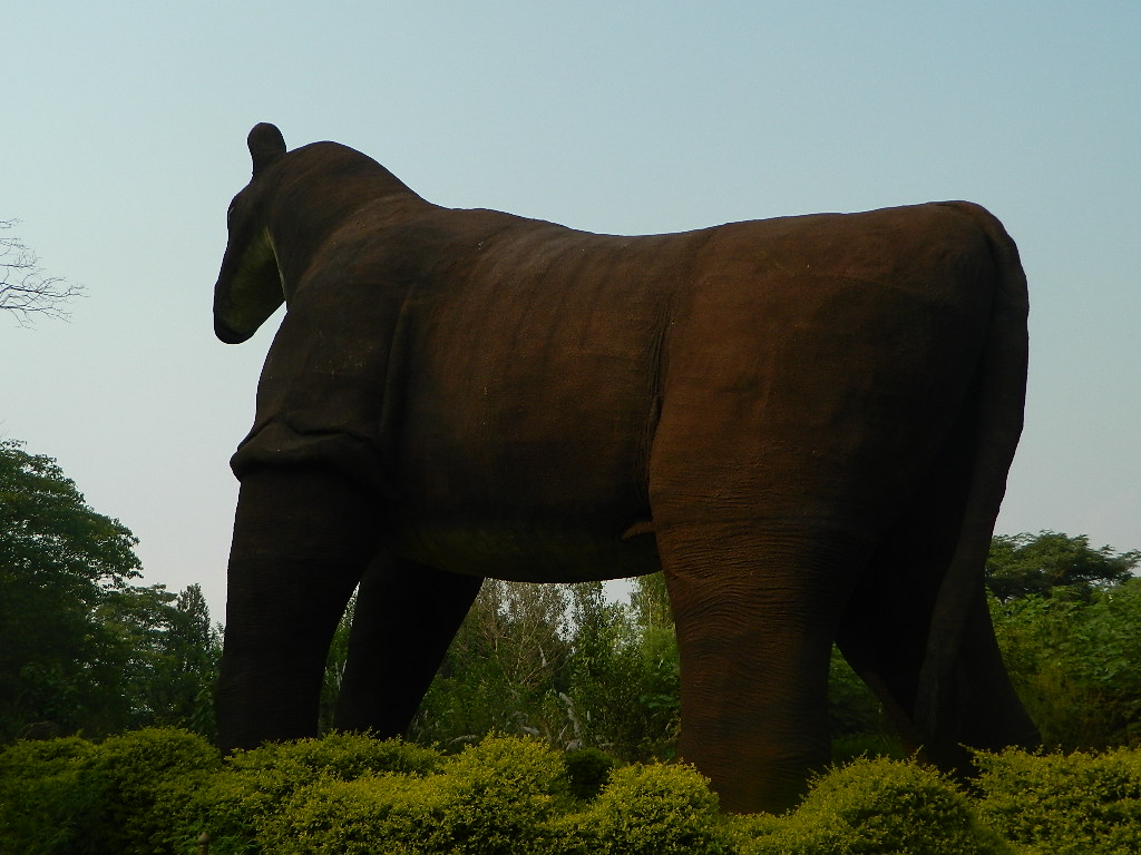 A large, brown sculpture of a horse stands against a blue sky, surrounded by lush green bushes and trees. The horse faces away, creating a serene and majestic atmosphere.