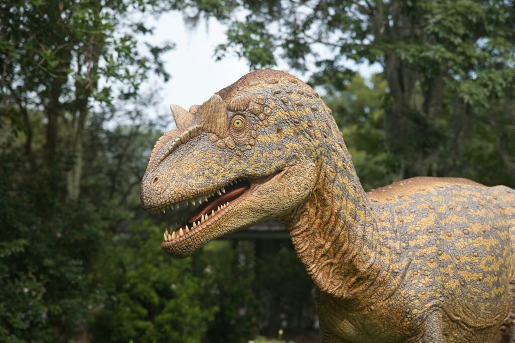 A lifelike dinosaur sculpture, possibly a carnivorous species, with textured scales and sharp teeth, set against a backdrop of lush green trees.