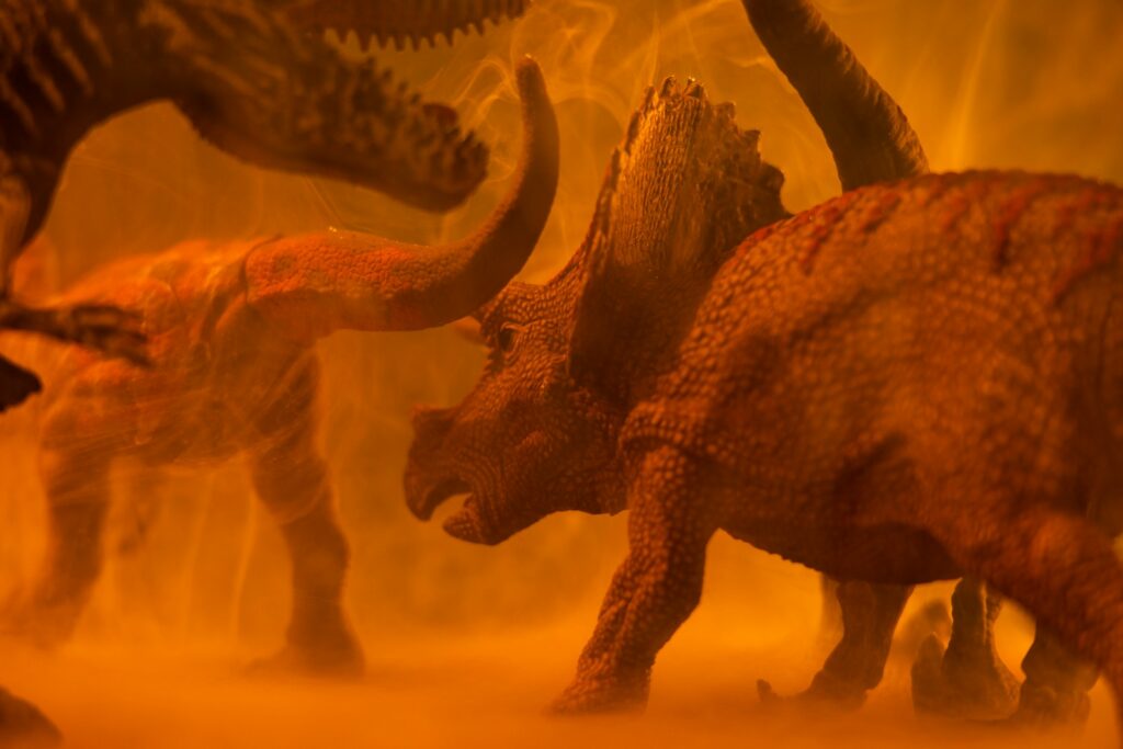 Figurines of dinosaurs, including a Triceratops, face each other amid swirling orange mist. The scene feels dramatic and tense.