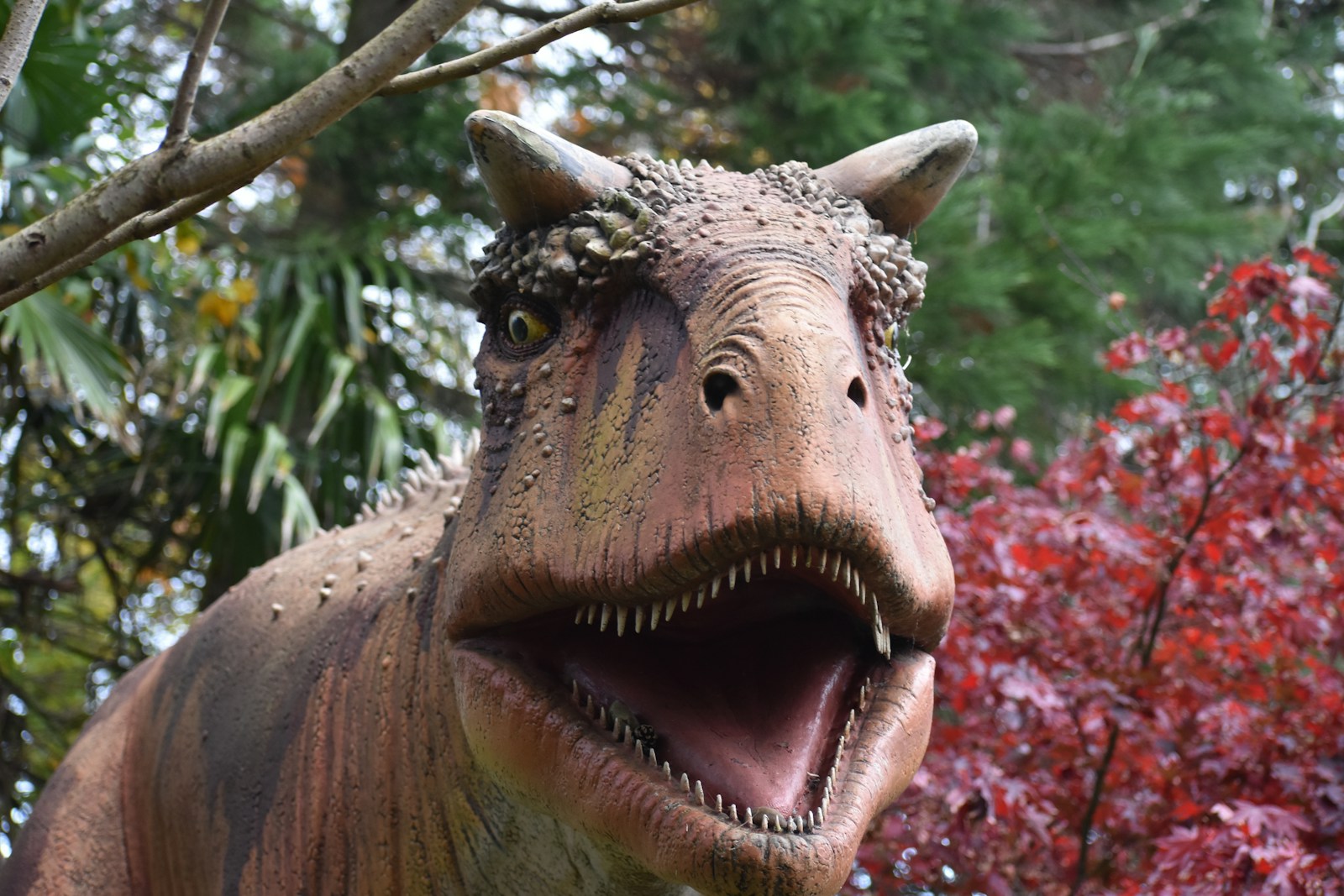 Dinosaur model in a garden setting with autumn foliage. Its mouth is open, showing teeth, creating a fierce expression. Green and red leaves surround it.