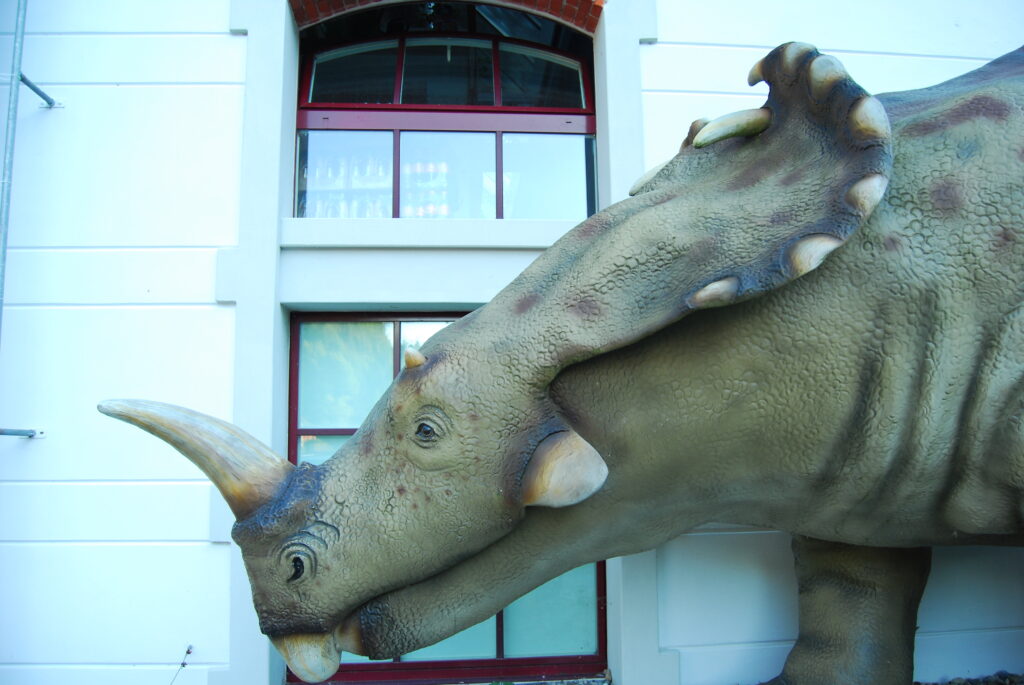 A detailed dinosaur sculpture, stands against a white building with red-framed windows, creating an intriguing prehistoric scene.
