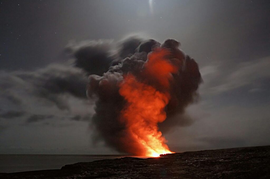 Molten lava glows red against a dark night sky, billowing smoke rising dramatically. The scene conveys power and intense natural energy.