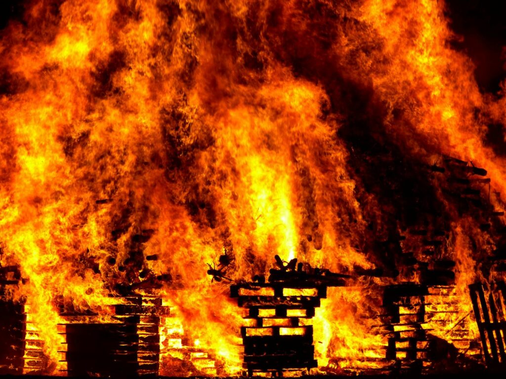 A massive, intense fire engulfs stacked wooden pallets, with bright orange and yellow flames roaring upward, conveying heat and destruction.