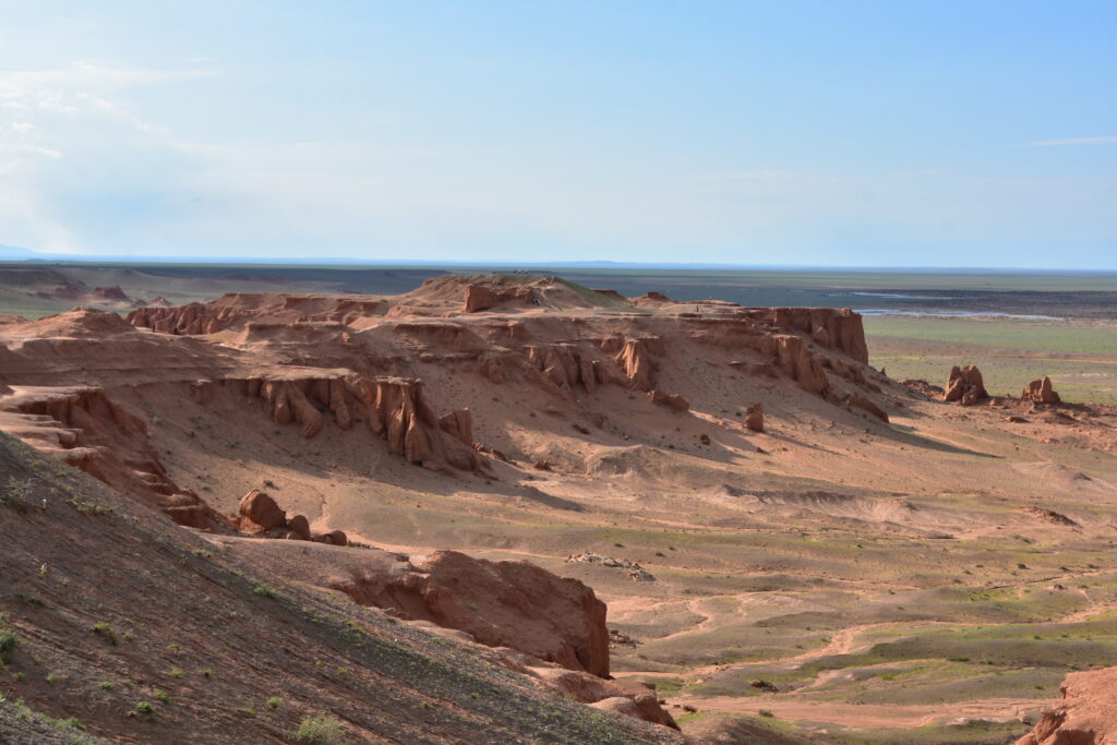 Desert landscape with layered, eroded hills in shades of pink and gray under a clear blue sky. Rugged mountains are visible in the distant background.