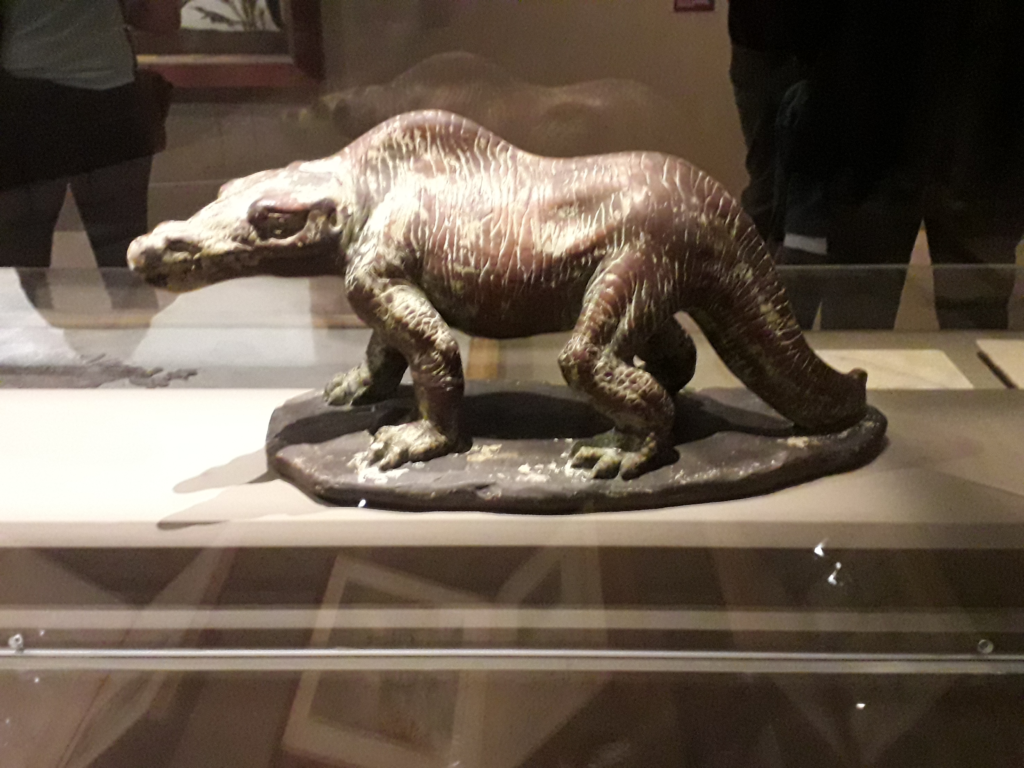 A small brown animal sculpture resembling a prehistoric creature is displayed in a glass case. It stands on a platform with textured skin detail.