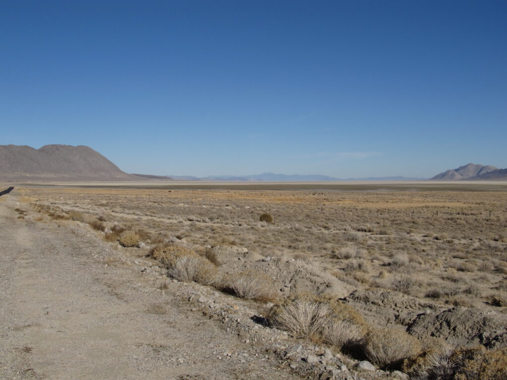 Arid landscape with a dry, flat plain stretching to distant mountains under a clear blue sky. Sparse shrubs cover the rocky, beige terrain.