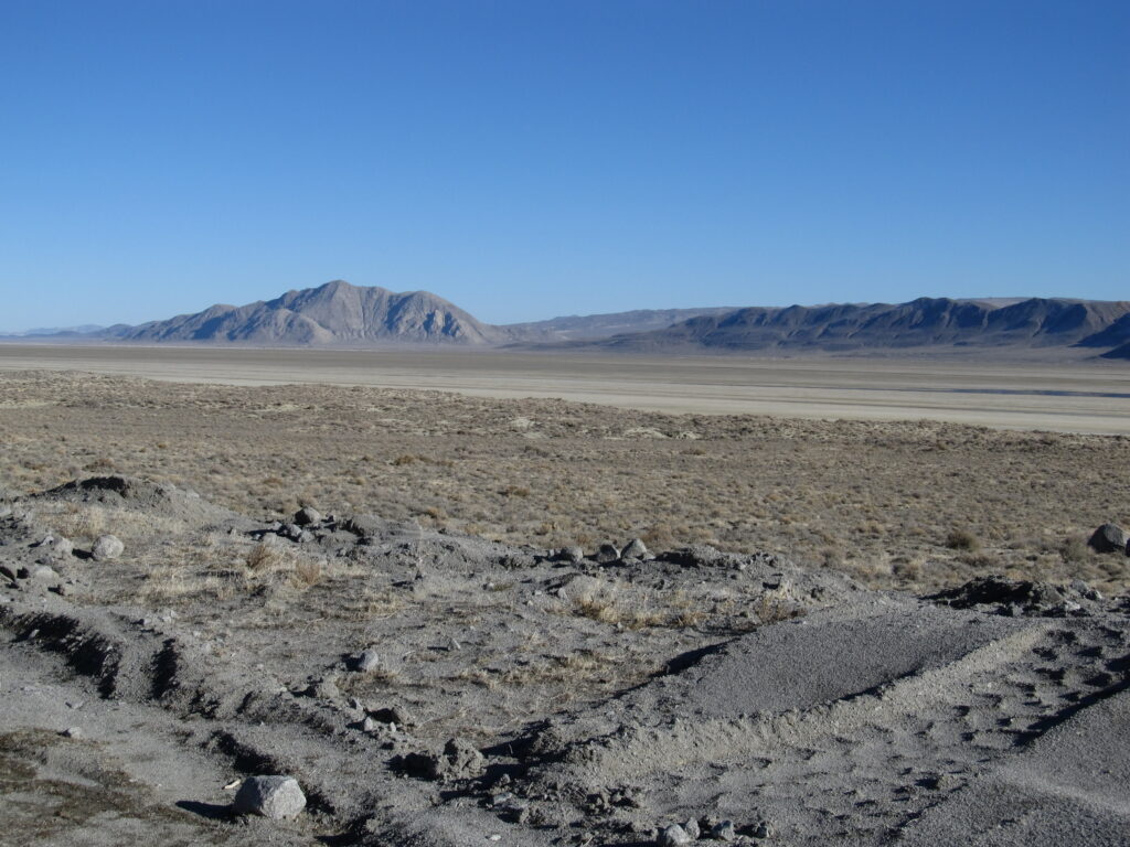 Arid landscape featuring a foreground of rocky, sparse terrain and tire tracks, with a flat, expansive desert plain leading to distant towering mountains. Sky is clear.