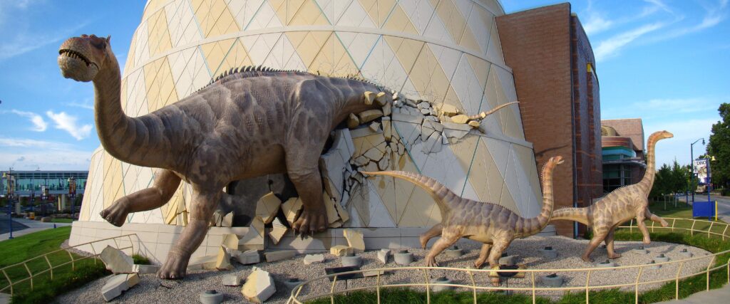 Giant dinosaur sculptures breaking through a wall at a museum. The scene is dynamic and dramatic, capturing a prehistoric theme with vibrant detail.