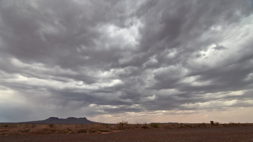 Dramatic overcast sky filled with dark, heavy clouds looming over a flat, arid landscape. The scene conveys a tense, moody atmosphere.