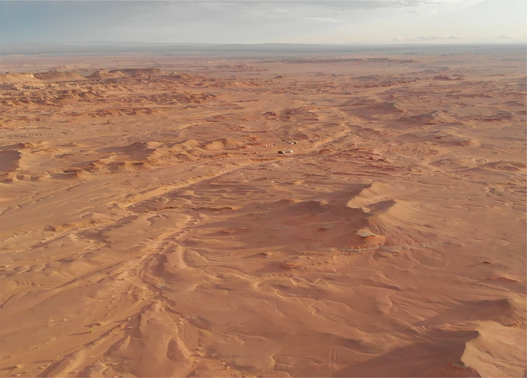A vast, arid desert landscape under a cloudy sky, with rolling dunes and small rugged hills. Sparse vegetation, conveying a sense of isolation.