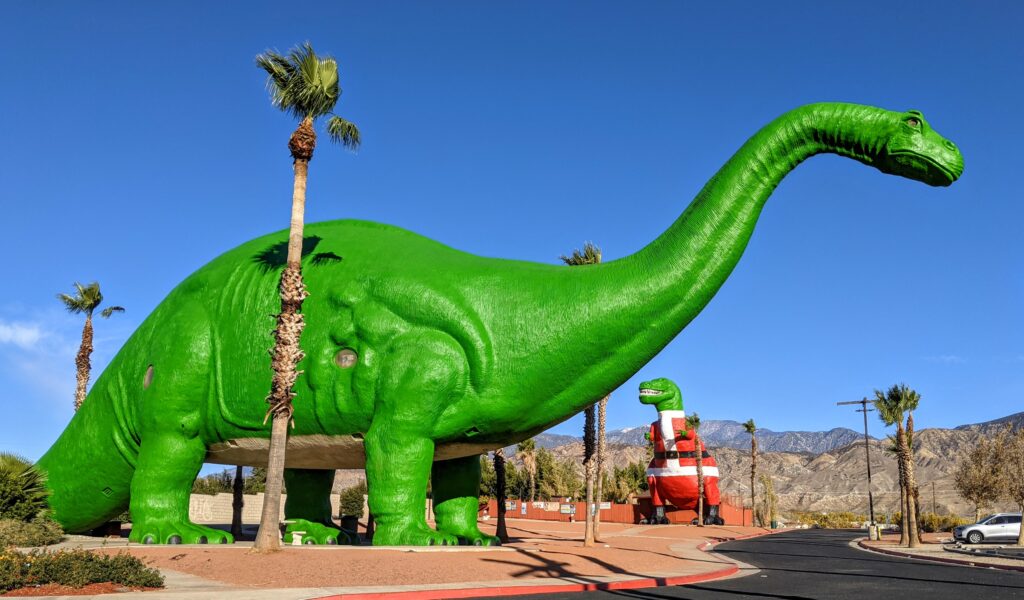 Large green dinosaur sculpture set against a clear blue sky, surrounded by palm trees. A smaller red dinosaur is visible in the background. Playful and whimsical tone.