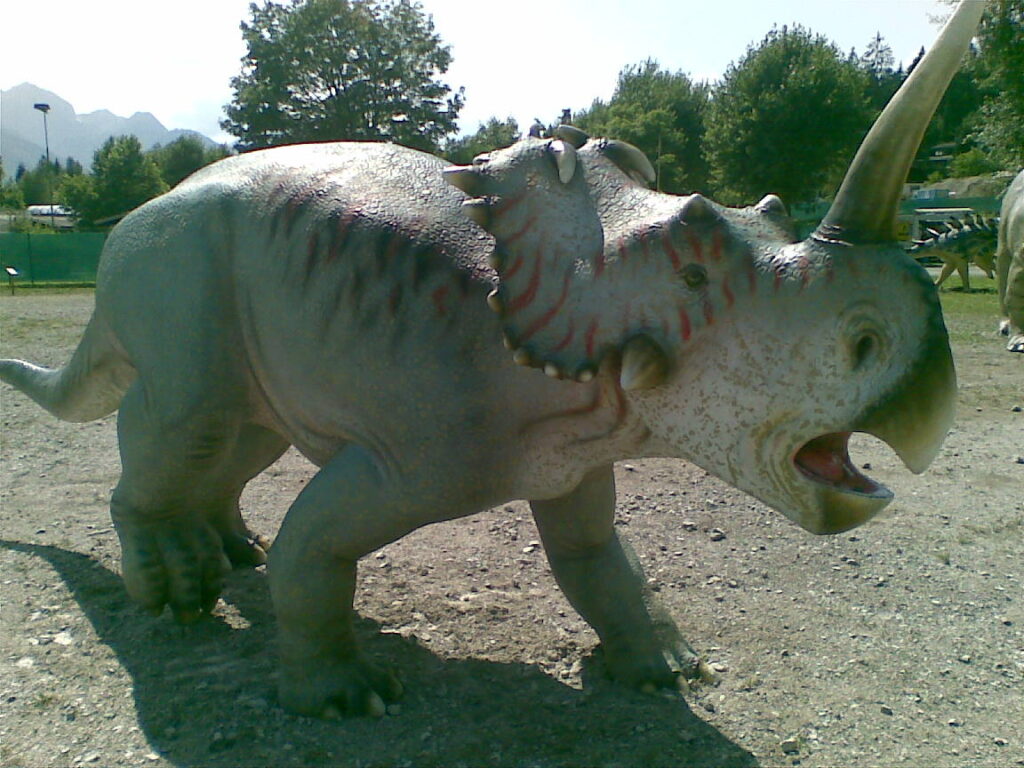 A large dinosaur statue with a horn and open mouth stands on gravel. It's outdoors, surrounded by trees under a sunny sky, conveying a prehistoric theme.