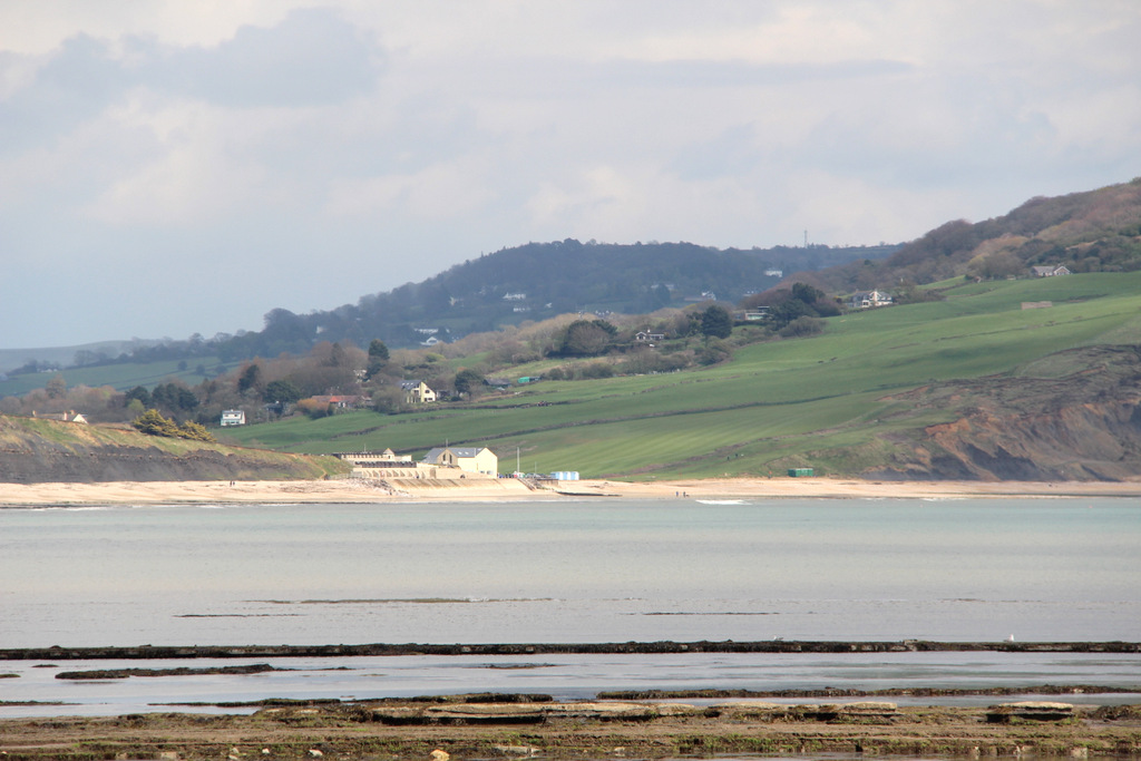 Coastal landscape with a sandy beach in the foreground and rolling green hills in the background. A few houses are nestled on the hills, under an overcast sky.