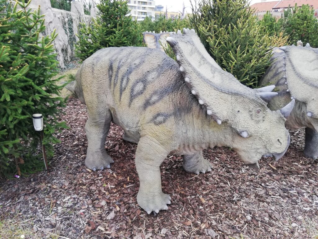 A realistic dinosaur statue with a textured, gray and brown body and prominent horns stands on mulch, surrounded by green shrubs and pine trees.