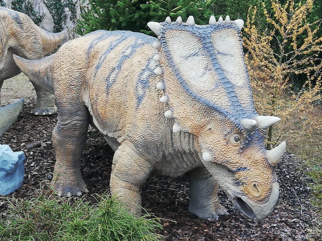 Model of a horned dinosaur with textured, beige scales and blue markings, standing on mulch. The background features greenery and small trees.