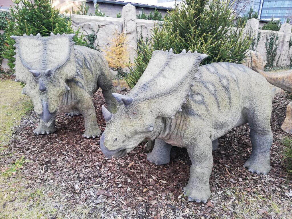 Two realistic dinosaur sculptures, stand in a garden with mulch and greenery. Their detailed textures suggest a prehistoric scene.