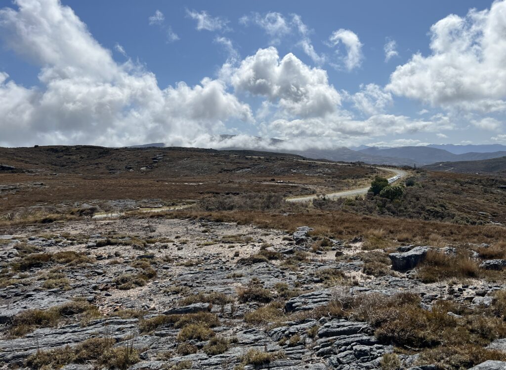 Rocky terrain with sparse vegetation under a bright blue sky, scattered with white clouds. Distant hills and a winding road add depth and tranquility.