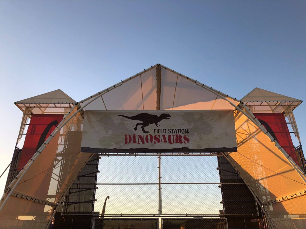 Entrance to "Field Station: Dinosaurs" with a canopy structure and dinosaur silhouette banner, against a clear blue sky during sunset.