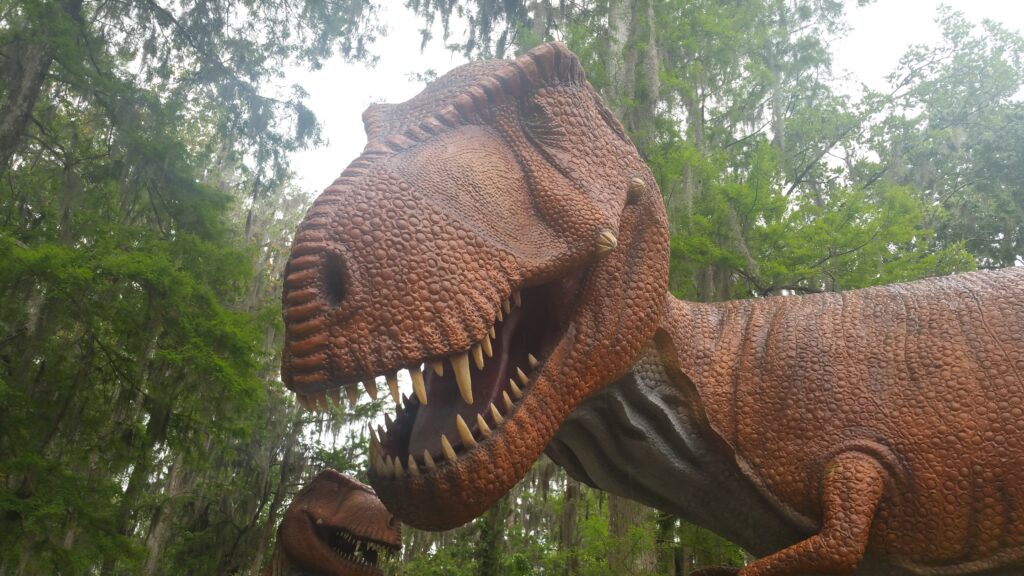 A large, brown dinosaur statue with textured skin and an open mouth showing sharp teeth is set against a backdrop of tall, green trees.