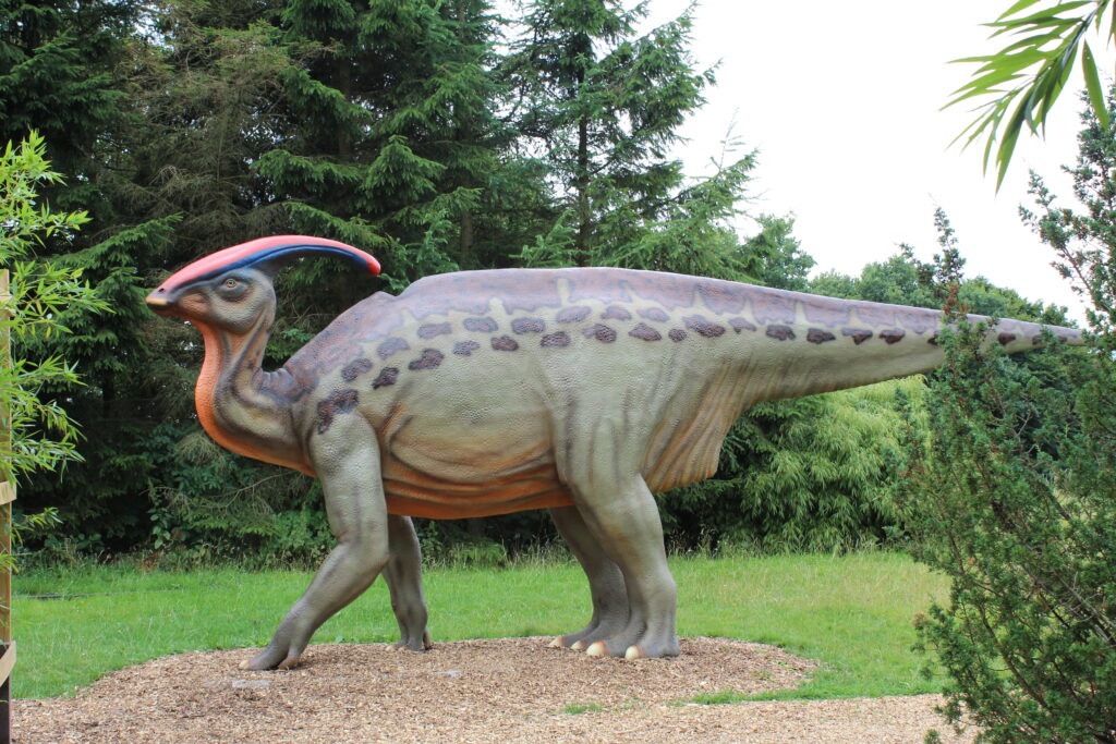 A life-sized statue of a Parasaurolophus dinosaur stands on wood chips, surrounded by lush greenery and trees, conveying a prehistoric feel.