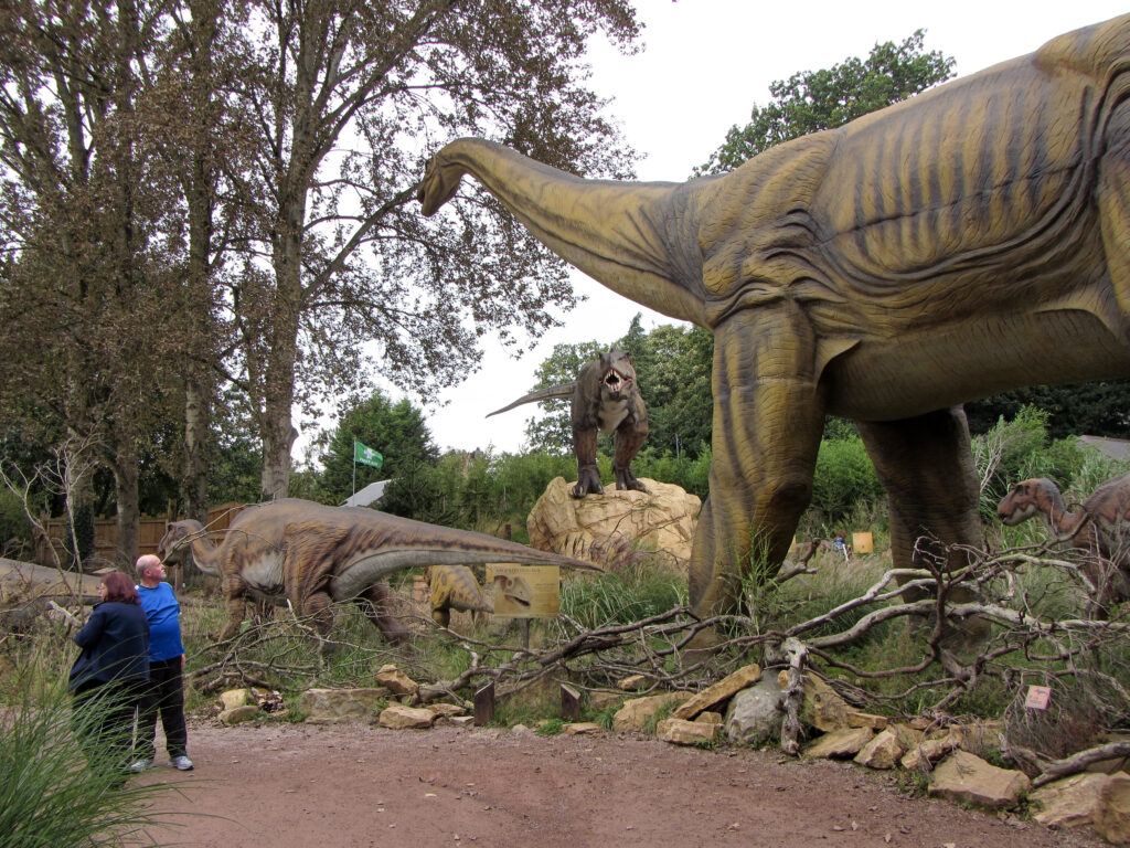 People look at life-sized dinosaur statues in a lush, outdoor setting. A towering sauropod and fierce theropod create a sense of awe and excitement.