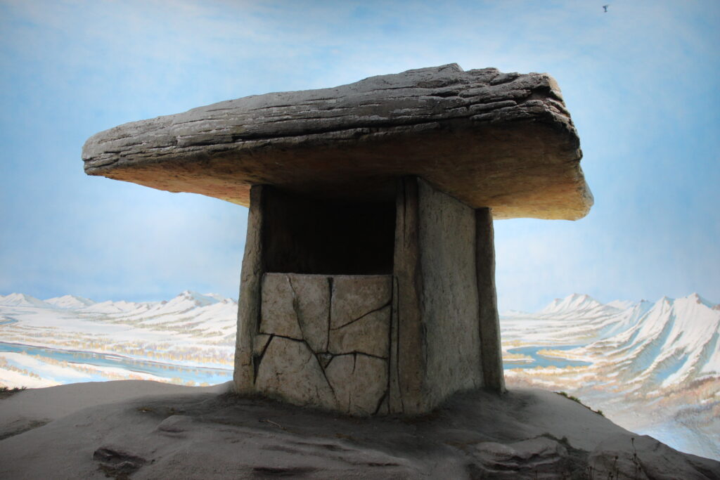 Stone dolmen against a backdrop of snowy mountains and blue sky. The structure is massive with a flat top, conveying a sense of ancient mystery.