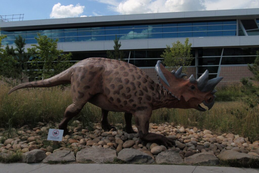 Dinosaur statue with horns and a speckled pattern stands on rocks in front of a modern building, surrounded by grass and small trees, under a cloudy sky.
