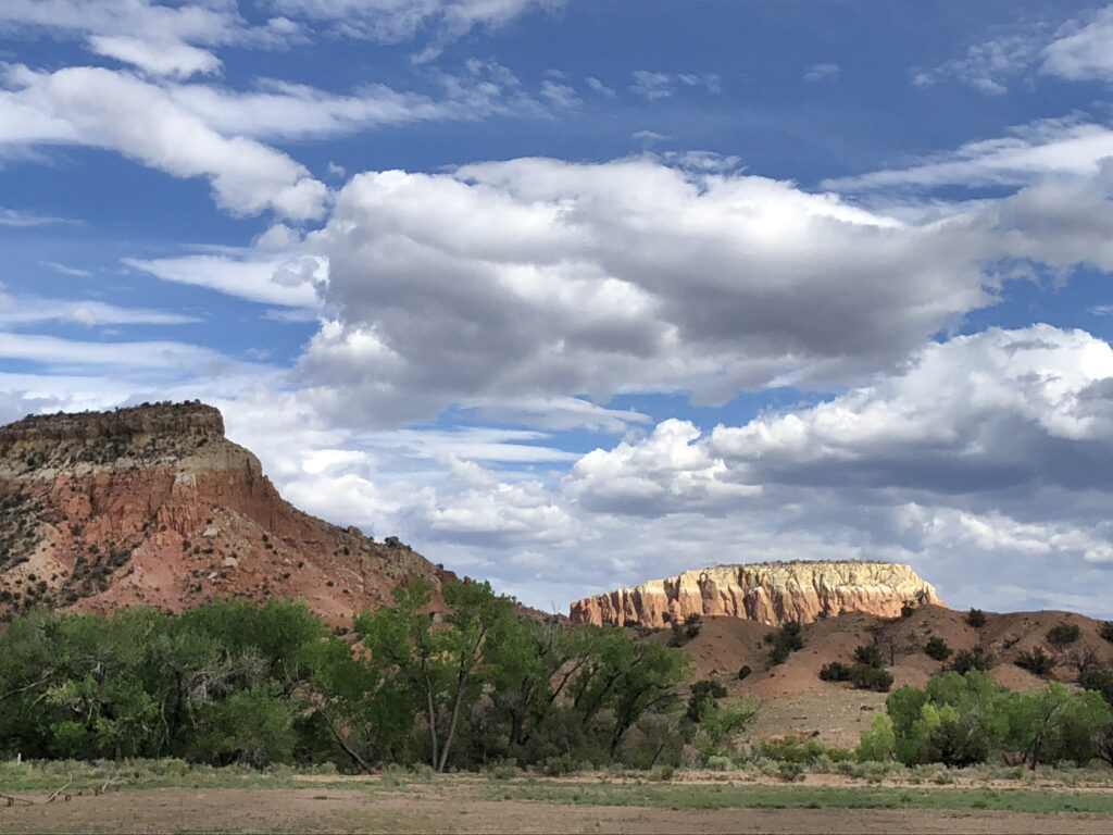 Expansive landscape with red rock formations and green trees beneath a vibrant blue sky filled with dramatic white clouds. Calm and serene scene.