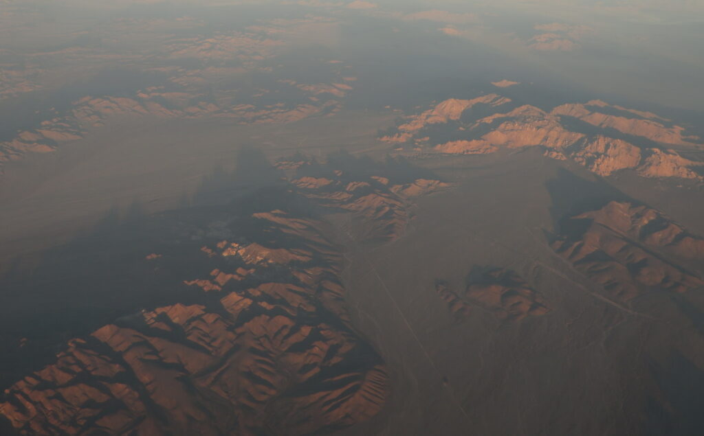 Aerial view of a vast desert landscape with rugged hills casting long shadows. The warm, golden hue of the setting sun highlights the terrain.