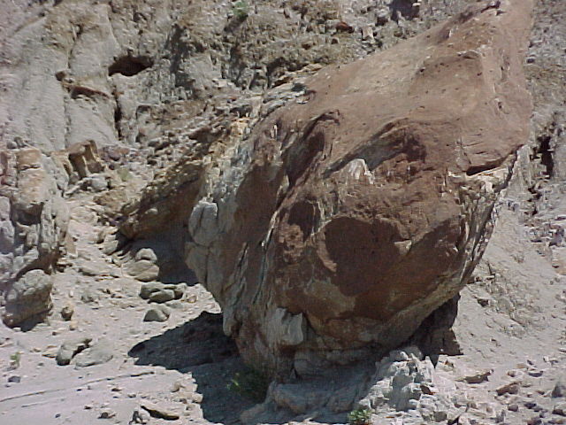 A large, reddish-brown boulder sits at the base of a rocky cliff, surrounded by smaller stones. The scene conveys a rugged, natural landscape.