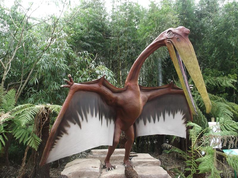 A large, realistic Quetzalcoatlus model with wide open wings and a long beak stands on a rock surrounded by lush greenery and ferns in a forest setting.