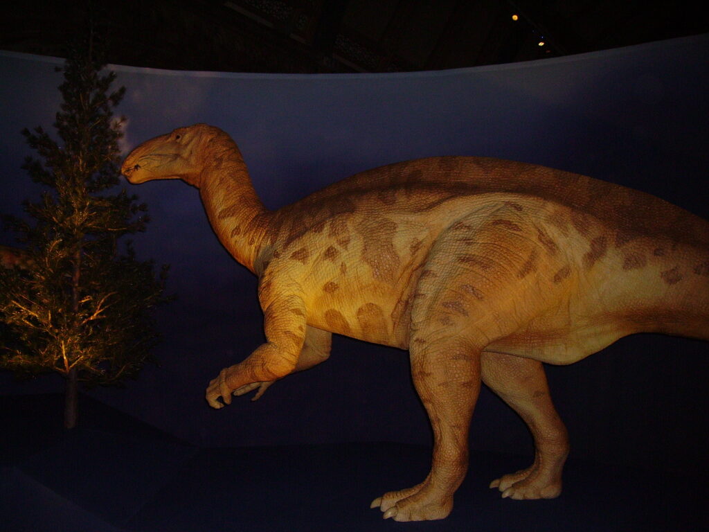 A lifelike dinosaur exhibit features a large, brown herbivore standing near a tree in a dimly lit museum setting, creating a prehistoric atmosphere.