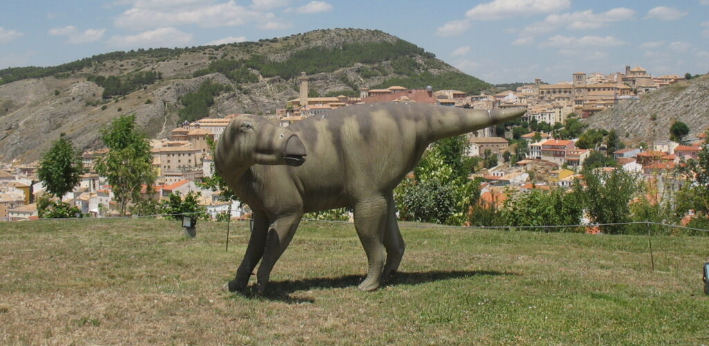 A dinosaur sculpture stands on grass with a mountainous village in the background. The sky is clear with a few clouds, creating a serene atmosphere.