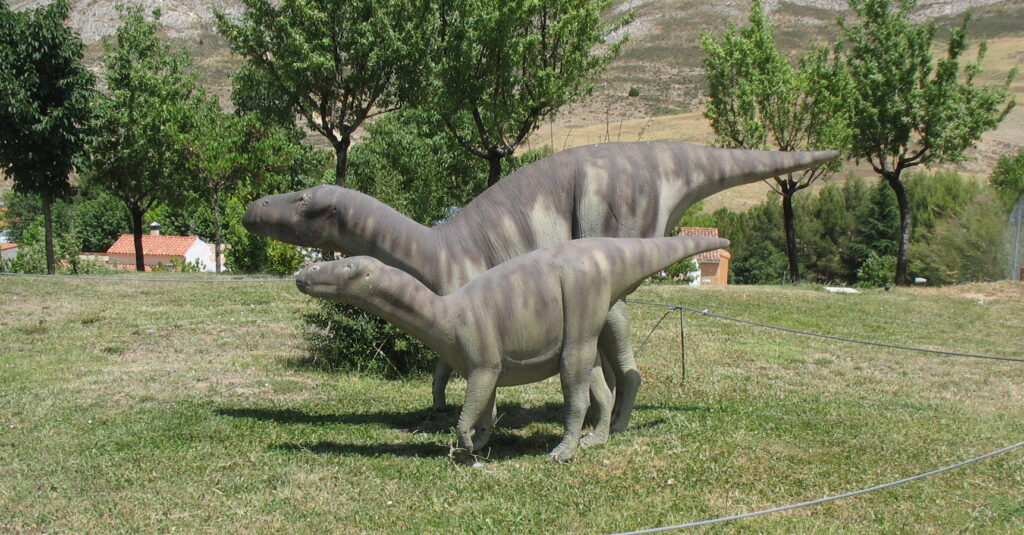 Two dinosaur sculptures stand on grassy terrain, surrounded by trees under a clear sky. The setting feels calm and natural.