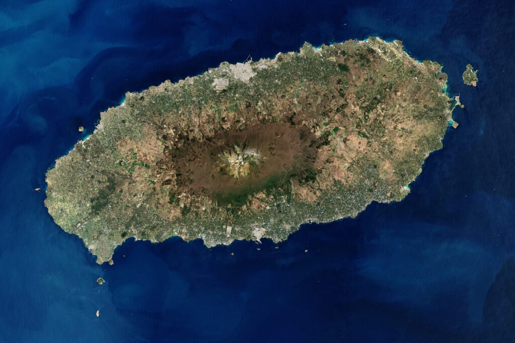 Satellite view of an island with a prominent central volcano surrounded by green and brown terrain. The island is encircled by deep blue ocean waters.