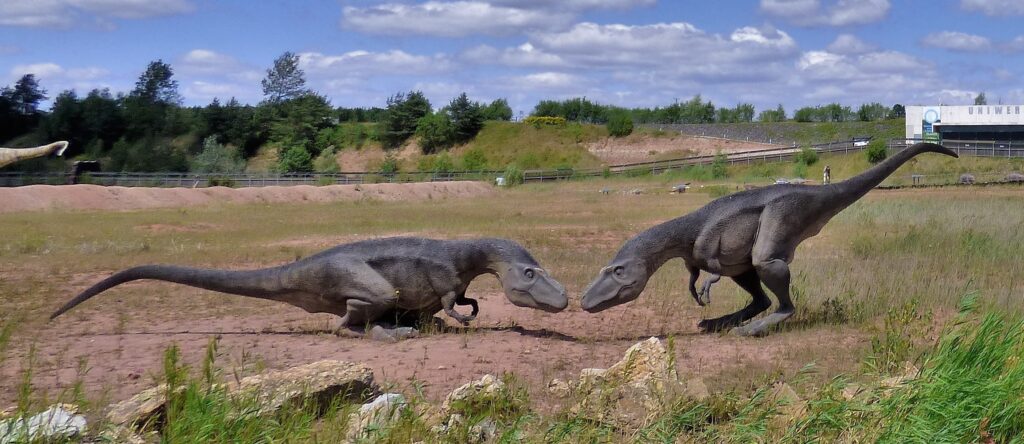 Two realistic dinosaur sculptures face each other in a grassy field under a blue sky with clouds, creating a sense of prehistoric interaction.