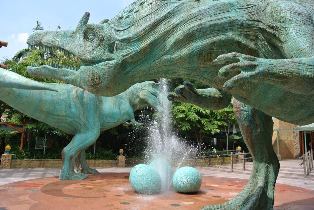 Large bronze dinosaur sculptures tower over a fountain with water spraying from dinosaur eggs. The lush greenery creates a prehistoric ambiance.