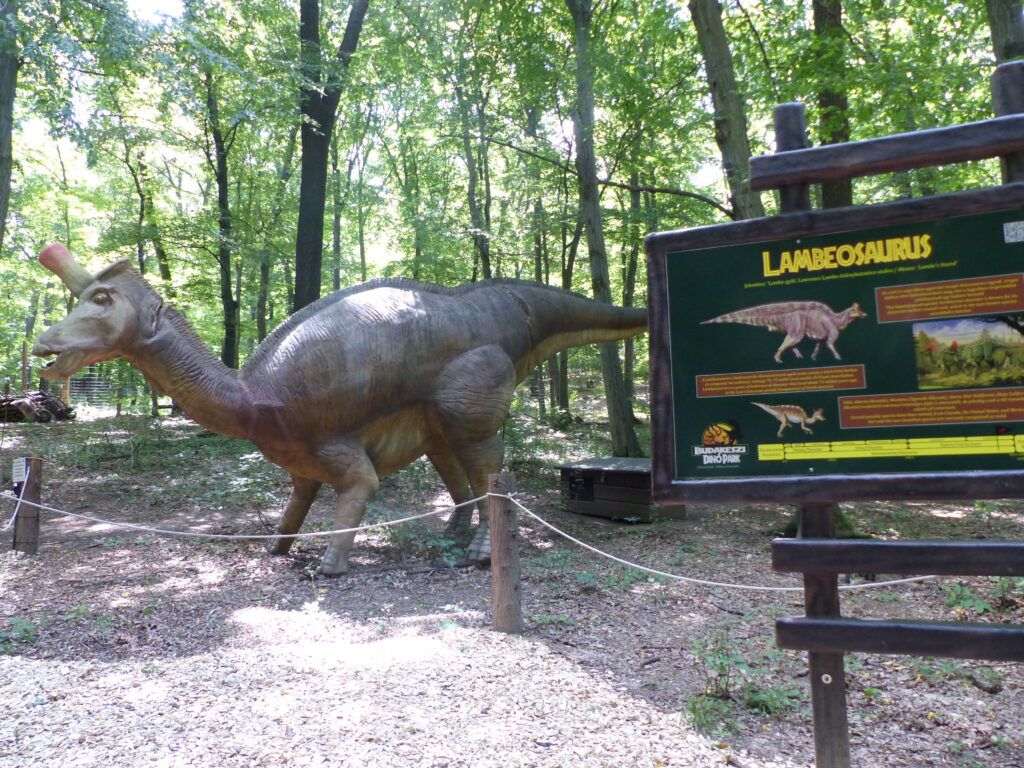 A life-sized Lambeosaurus dinosaur model stands in a wooded park. Sunlight filters through the trees. An informative sign to the right details the species.