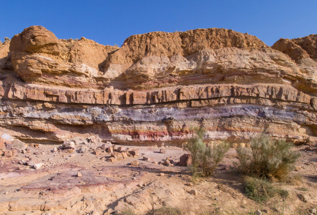 Rocky cliff face with layered strata in hues of red, pink, and beige under a clear blue sky. Sparse vegetation grows at the base, conveying an arid atmosphere.