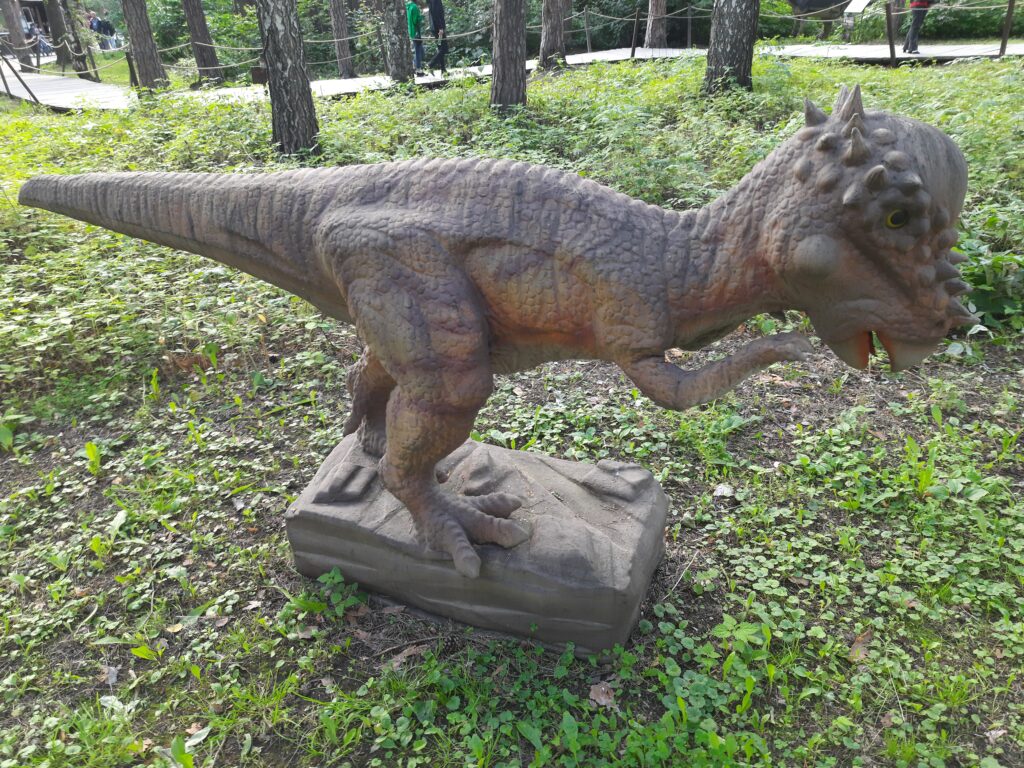 A brown dinosaur statue with a textured body and small spikes stands on a stone base, surrounded by greenery and trees, in an outdoor setting.