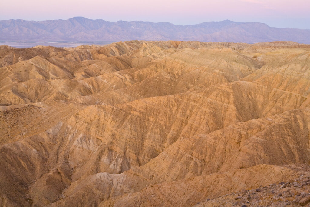 Vast desert landscape with rugged, sunlit brown hills and ridges, framed by distant mountains under a soft pink sky, creating a serene atmosphere.